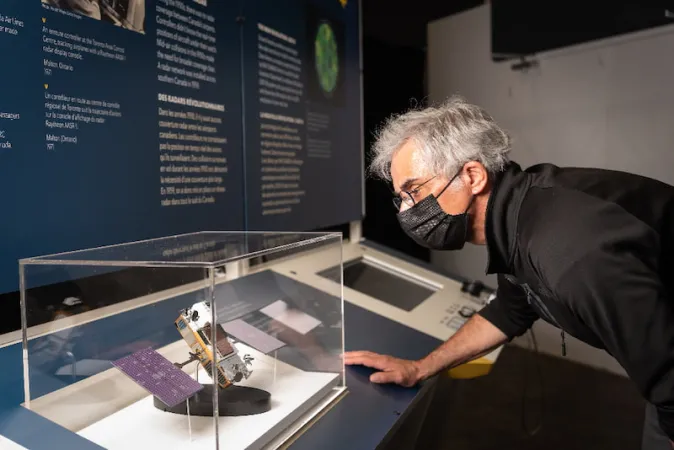 A silver-haired man leans in to look at an encased model of a satellite in front of one of the exhibition modules.