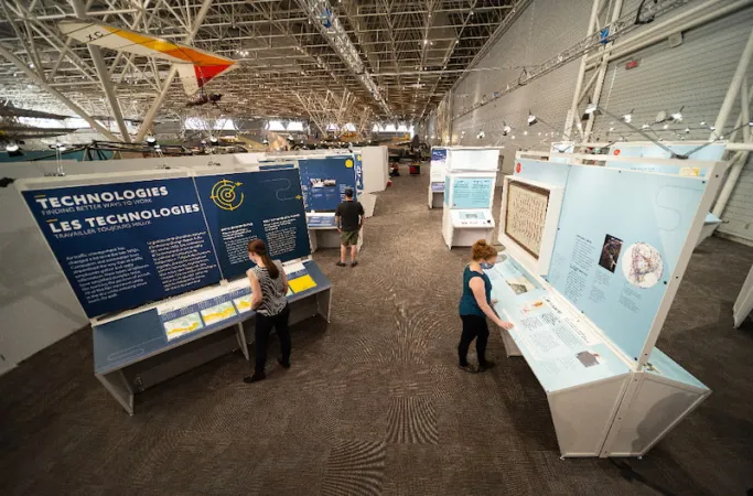 An aerial view of the exhibition, showing many modules with three visitors throughout. In the foreground, the word “Technologies” is visible in large letters on one of the panels.