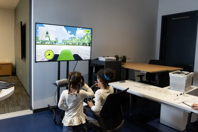 Two kids with curly hair in a bun and in braids looking at a computer screen featuring the simulation of a tractor. One of the kids is sitting down holding a steering wheel.