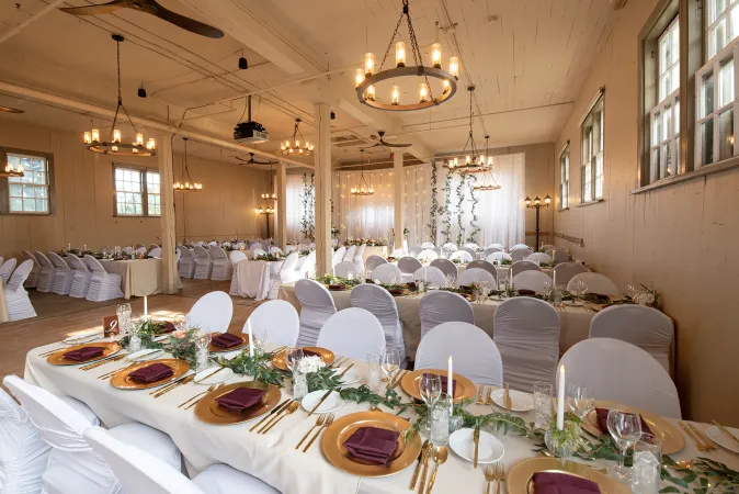 A beautifully decorated room with high white ceilings, white exposed brick walls and wood flooring. Multiple tables are lined up around the room draped in white table cloth with white fabric chairs. Golden plates and mahogany napkins are lined up at the tables with white candles.