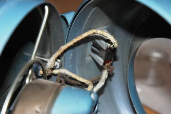 L’intérieur d’un ventilateur révèle des fils électriques dont la gaine d’amiante est exposée. Les fils, beige et blanc, s’effilochent. 