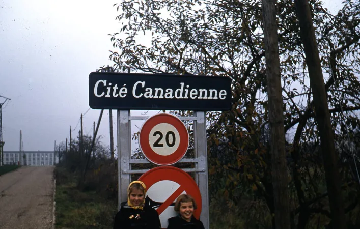 Two young children stand outdoors under a sign saying 'Cite Canadienne' and the number 20 in a red circle