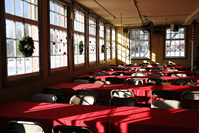 Tables lined up in rows in a room with large windows along one side of the room. Red tablecloths are draped over the tables and dark foldout chairs line each of the tables.