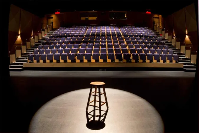 The view from a stage of a large theatre. Rows of purple chairs on an incline face the stage where there is a stool set up centre stage with a spotlight shining down on it. Two sets of stairs lead up the rows of chairs to the back where there are two red exit signs hanging over two doors.
