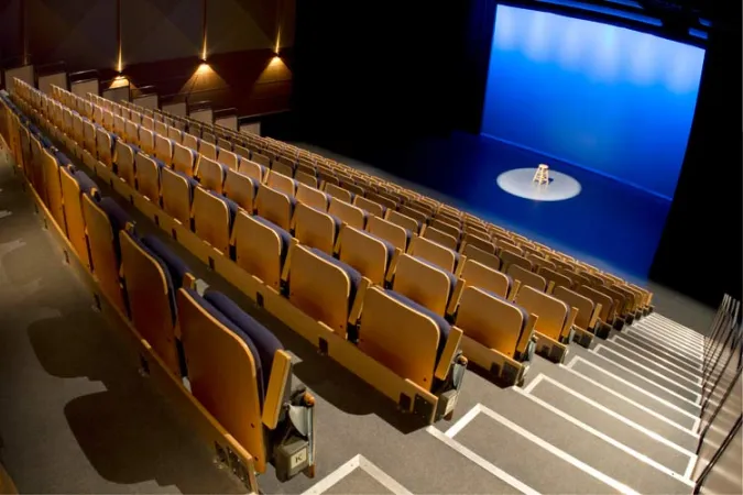 Large theatre with the view looking down onto a stage from the back of the room. Blue curtains line the stage and the background of the stage is lit in blue with a stool in the spotlight in the centre of the stage.