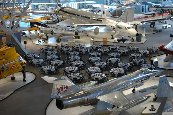 A bird’s eye image of an indoor scene with dozens of large round tables set up in between several airplanes in the centre of a room. The tables are draped with white table cloths and the chairs are a dark blue colour. Multiple different coloured airplanes surround the tables.