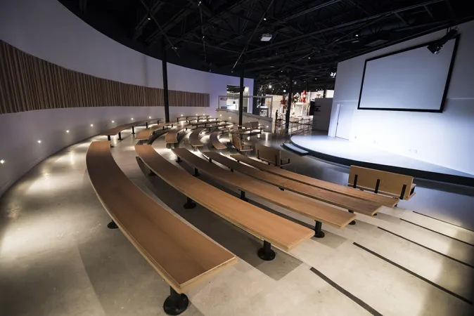 L’espace Curiosité en scène au Musée des sciences et de la technologie du Canada. La salle comporte une scène en demi-cercle avec un grand écran. Des rangées de bancs en bois de forme incurvée entourent la scène et plusieurs escaliers mènent à la scène. D’autres options de sièges sont offertes à l’avant, près des escaliers.