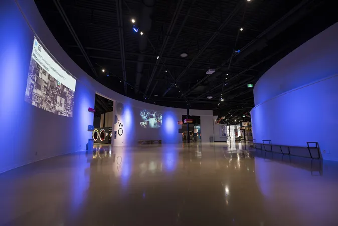 Une grande pièce vide au milieu du Musée des sciences et de la technologie du Canada. Les murs sont courbés et blancs, le plafond est haut et peint en noir, avec des projecteurs bleus et blancs qui diffusent une lumière tamisée. Une diapositive est projetée sur un mur.