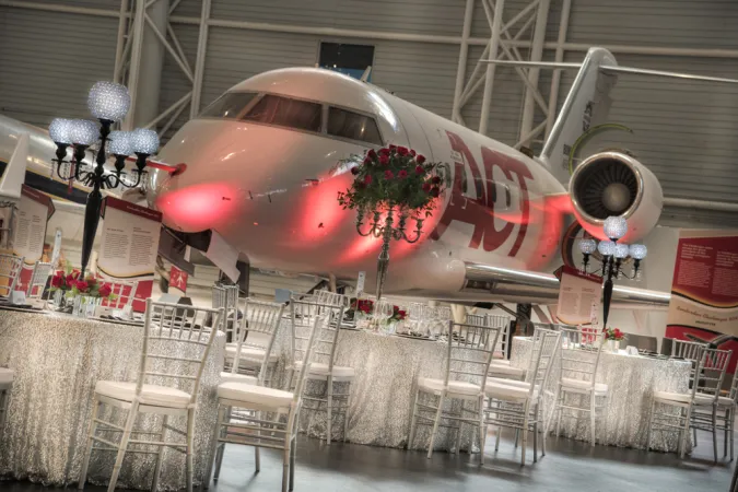 A view inside the Canada Aviation and Space Museum with a large white airplane in the background. Large tables are set up around the room with white translucent shimmering table cloths and elegant white chairs. Scattered around the room are classical Victorian-style lampposts with round lamp bulbs with geometric patterns on the glass