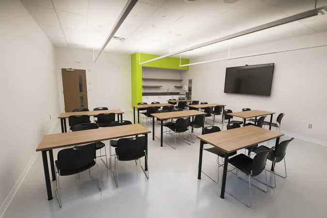  A spacious classroom with a TV on one side and eight desks with four chairs each arranged around the room. Another wall has a sink and a few shelves.