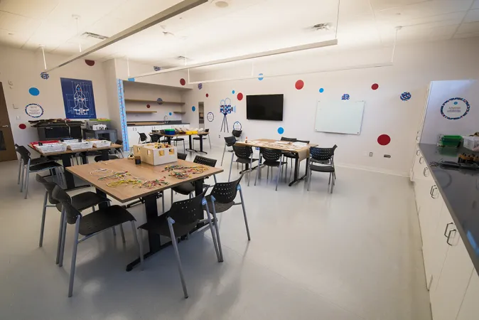 A spacious classroom with eight tables arranged in groups of two with five to six chairs at each table. A TV is displayed on one side of the room and the walls are decorated with colourful patterns including red and blue polka dots, gears, and the outline of a videocamera. Cabinets line the floor along one wall.