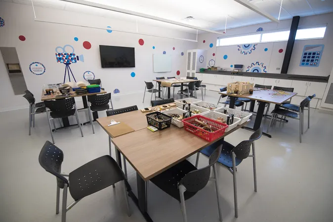  A spacious classroom with eight tables arranged in groups of two with five to six chairs at each table. A TV is displayed on one side of the room and the walls are decorated with colourful patterns including red and blue polka dots, gears, and the outline of a videocamera.