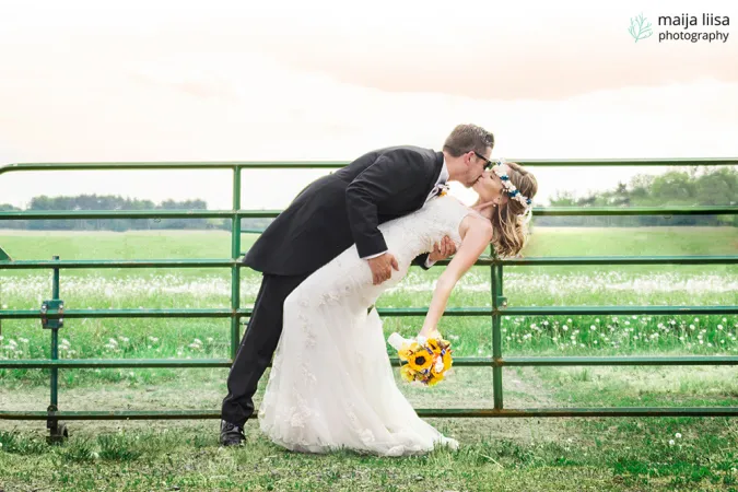 A groom tips a bride backwards as he kisses her in front of a green steel fence. Through the fence, a large green pasture with trees and a bright sky can be seen in the background.