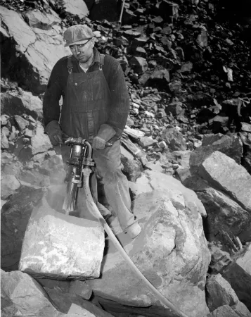 A man breaking large blocks of iron ore with an air drill, blasted from the face of a hill of ore in Eastern Canada so it will pass through the crusher.  