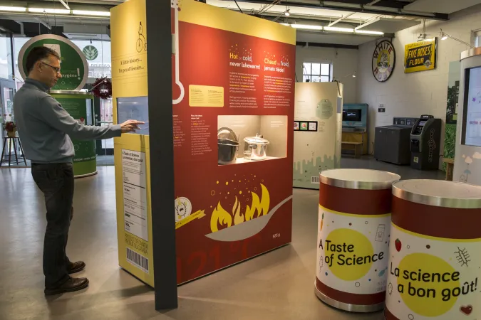 A person is standing in front of a touch screen that is part of an exhibition module.  The module also has a display case showing canning pots. Two small cylinders are to the right with the words “Taste of Science.”