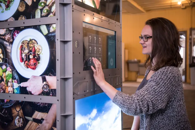 A museum visitor touches a screen full of question marks. Around the corner on the same exhibition module, plates of food are displayed.
