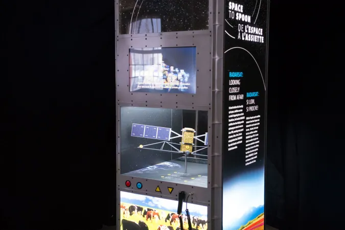 Un module d’exposition rétroéclairé, sur fond noir. On y voit une maquette de satellite, et le titre de l’exposition, « De l’espace à l’assiette ».