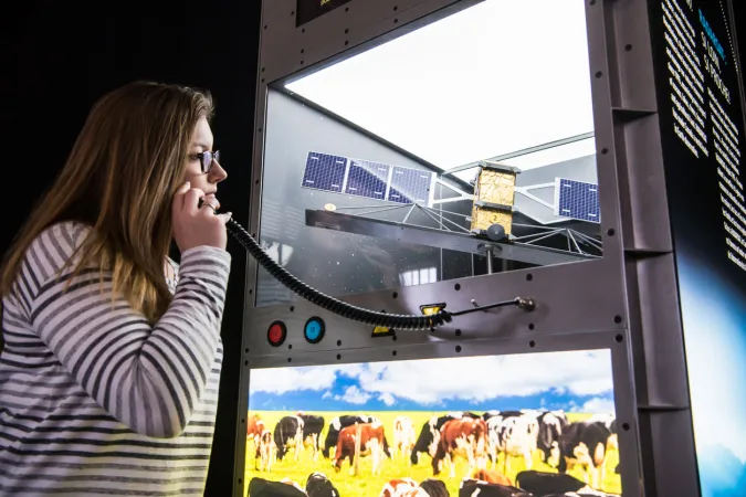 A museum visitor holds a listening device to their ear. The exhibition module in front of them contains a model of a satellite and an illuminated photograph of cows in a field.