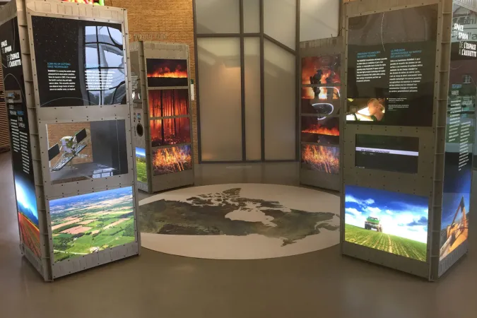 Dans une pièce lumineuse, quatre modules d’exposition rétroéclairés en forme de tour, disposés autour d’une carte du Canada posée au sol.