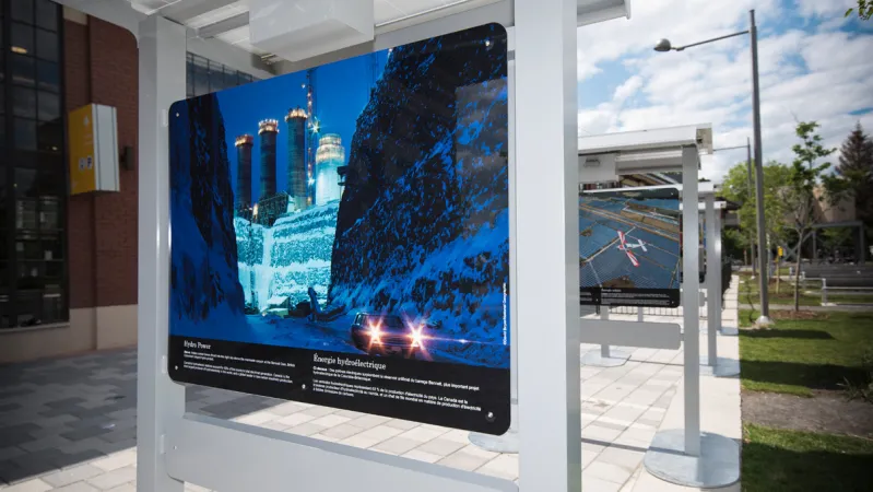 Les panneaux d’une exposition photographique sont installés en cascade, à l’extérieur. La première photo est celle d’une centrale hydroélectrique en Colombie-Britannique, en hiver. Il y a du texte blanc sous la photo.