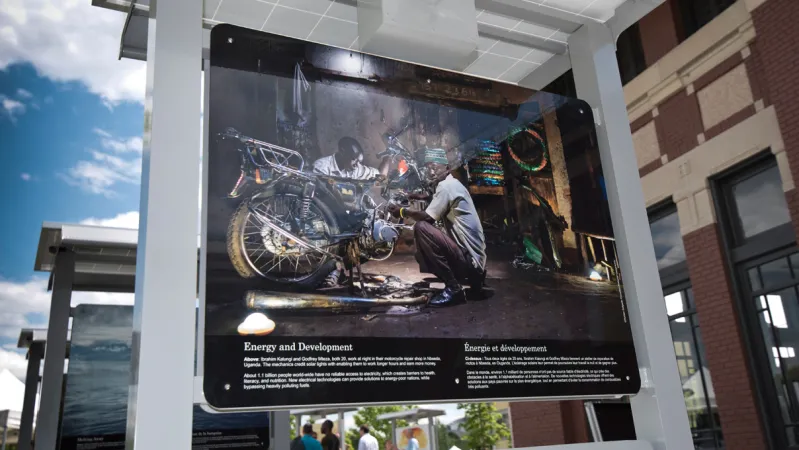 A panel from a photographic exhibition that shows a motor cycle repair shop with two people squatting next to a motorcycle. There is white text below the photograph.