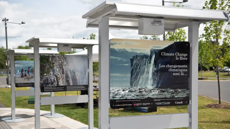 Three panels from a photographic exhibition are in a row, in an outdoor setting. The first photograph is of an iceberg with the title Climate Change is Here written in white text. The second photograph is of a cliff face, covered in mist.