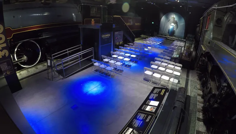 A top-down view of a large dark room lit with futuristic blue lights and a life-sized black and green train on one side of the room and a black and mahogany train on the other side of the room. On the other side of the wall is a large white cloth featuring the faint outlines of a steamboat. Several rows of white fold-out chairs are set up on one side of the room.