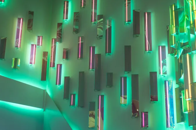 Gros plan sur les lumières dans l’entrée du Musée des sciences et de la technologie du Canada. Le mur est composé d’un ensemble de néons et de lampes fluorescentes ainsi que de miroirs disposés verticalement dans des rectangles de tailles différentes, créant un kaléidoscope de reflets et de couleurs. Sur la photo, les néons produisent des lignes magenta et le mur est rétroéclairé de teintes de vert, de jaune et de sarcelle.