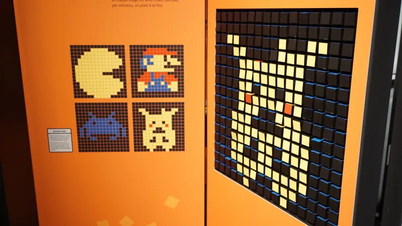 Two orange exhibition panels show an interactive game. The panel on the left has four squares with images of pixelated video game characters. On the right is an interactive game where the player turns squares to create a pixelated image.