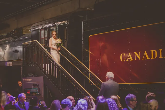 A bride holding flowers walking down the stairs with sleek black balusters and golden handrail that leads to the entrance of one of the trains at the Canada Science and Technology Museum. A person in a suit waits for her at the bottom of the stairs. The beginning of the word Canadian can be seen on the side of the train.