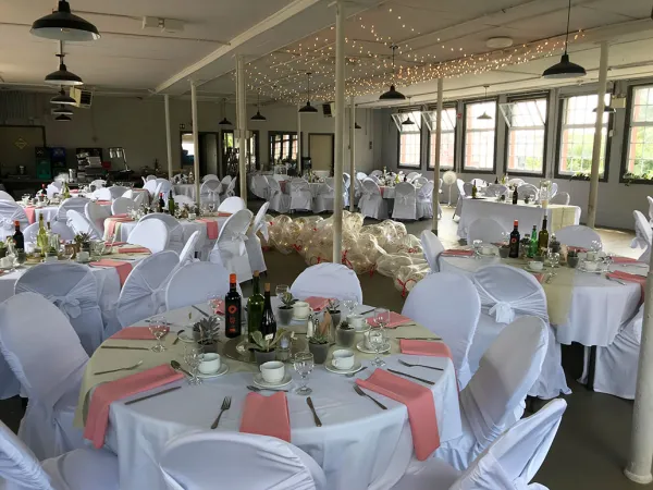 Room filled with several big round tables. Several chairs are placed around the tables and both are draped with floor length white tablecloths. The tables are decorated with silverware and pink napkins. String lights hang across the ceiling.
