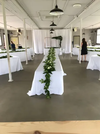 Une grande pièce au plafond blanc où sont aménagées plusieurs longues tables rectangulaires. Chaque table est drapée d’une nappe blanche et recouverte d’un chemin de table de verdure et de fleurs.