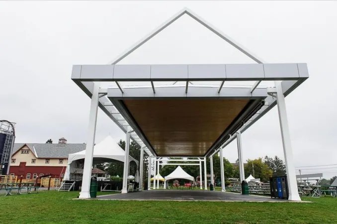  Outdoor pavilion with white roof and several tents set up in the background. Bleachers line one side of the pavilion and a barn can be seen in the background.