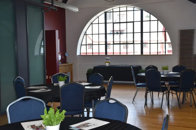 Une grande pièce avec des fenêtres en demi-cercle sur un mur et aux vitres multicolores sur un autre. Des tables rondes sont disposées dans la pièce, entourées de plusieurs chaises bleues.