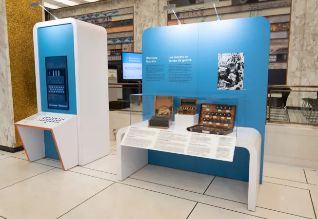 Two blue and white exhibition modules are side by each. The module to the left has a large screen.  The module to the right has a display case, with a enigma machine and cipher wheels on display. The panels behind the display case have black and white text and photo.