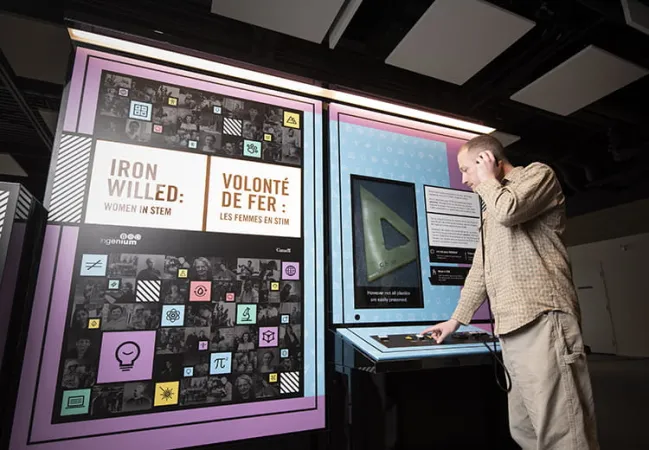 Un homme tient un écouteur contre son oreille et appuie sur un des boutons d’un comptoir interactif d’exposition. À gauche, un panneau porte le titre de l’exposition, « Volonté de fer : les femmes en STIM ».