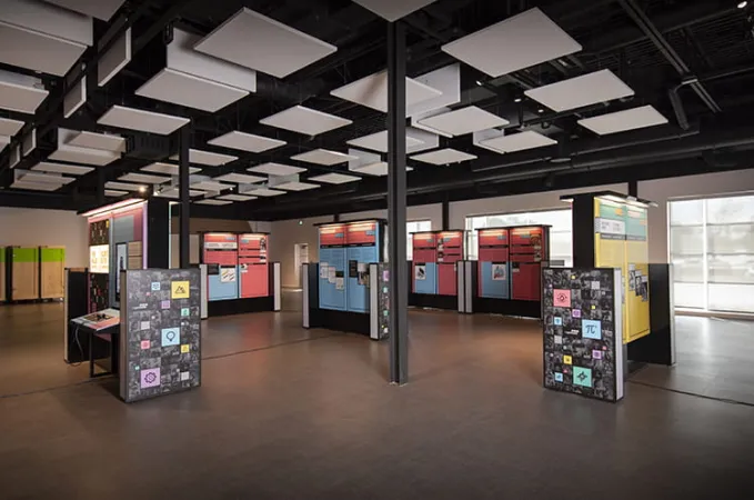 De nombreux modules d’exposition sont répartis dans une grande salle. Les panneaux sont de couleur vive, principalement du bleu, du rose et du jaune.