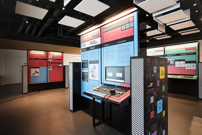 A view of three exhibition modules that are bright blue, pink, and green in colour. The module in the foreground includes an interactive with buttons and a TV screen.