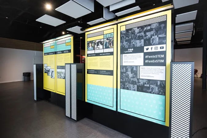 Two yellow and green exhibition modules are side-by-side. The hashtag “#FemInSTEM” is visible in the foreground.