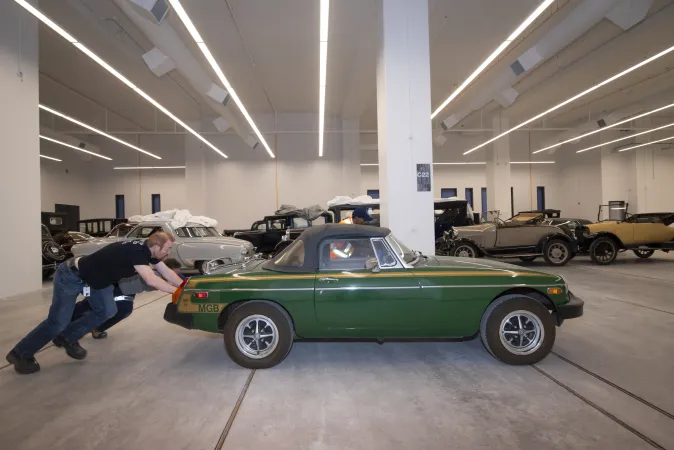 Two men push a green sports car next to a large collection of vehicles.
