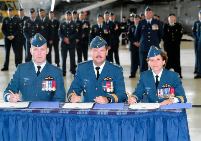 Three people in blue military uniforms sign documents at a table, while uniformed men stand behind them. 