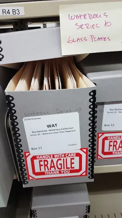 Photo showing an archival box containing glass plates with a large red 'Fragile' sticker on it