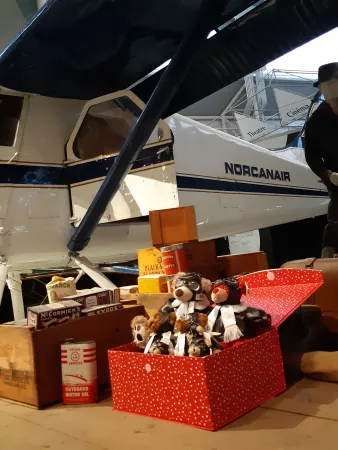 A red box filled with teddy bears dressed as pilots sits next to an aircraft in a museum.
