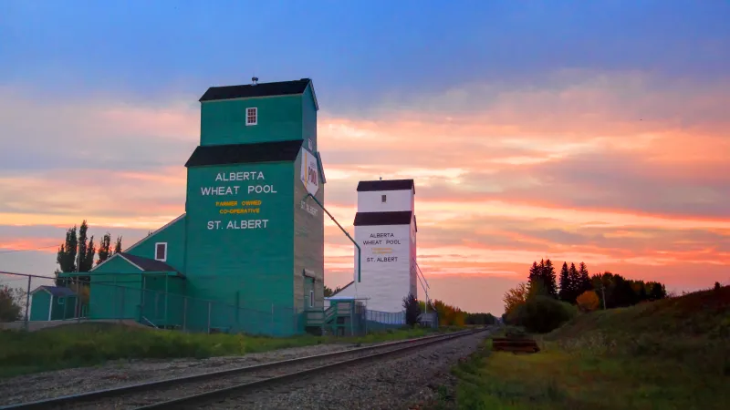 Two tall grain elevators stand alongside a railway track, with a beautiful sunset in the background.