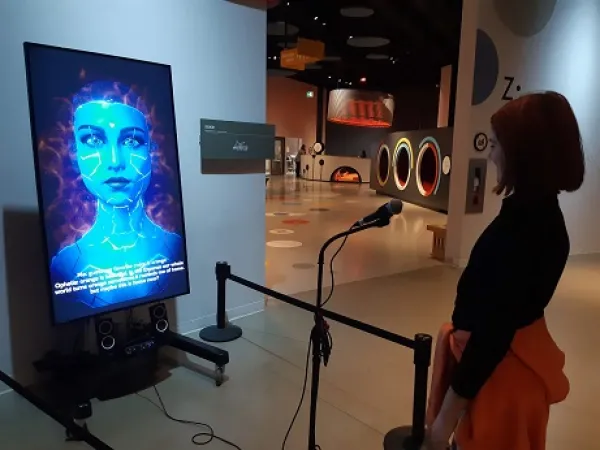Une jeune fille devant un microphone, regardant un grand écran affichant le visage d’un avatar féminin bleu. 