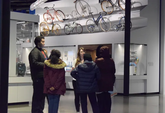 Un groupe d'adolescents et un coordinateur se réunissent pour faire le tri de leurs idées et trouver la solution. Derrière eux se trouve une vitrine qui présente les différents modèles de vélos ayant existé au fil du temps.
