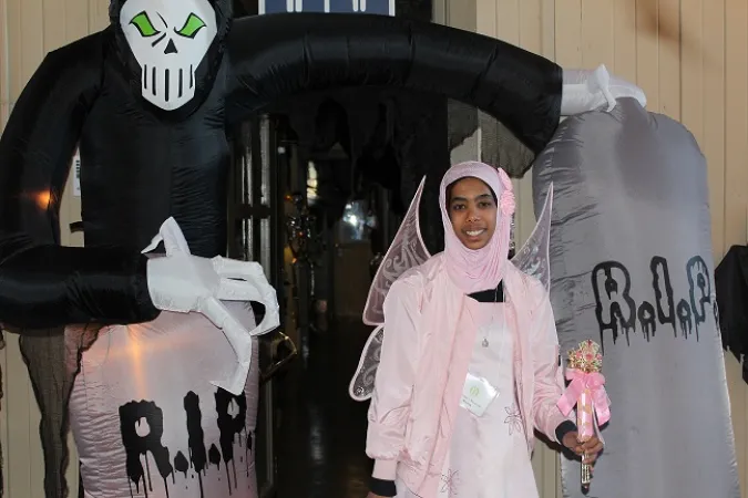 A young girl in a pink outfit and matching hijab wears a set of butterfly wings as she poses in front of a Halloween display.