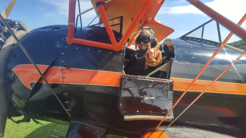 A man sits in the cockpit of a black and orange biplane.