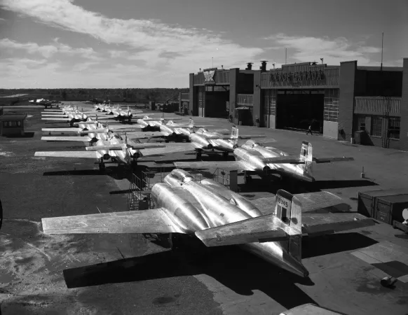 A black-and-white photograph showing a line of CF-100 jet aircraft in front of two hangars.