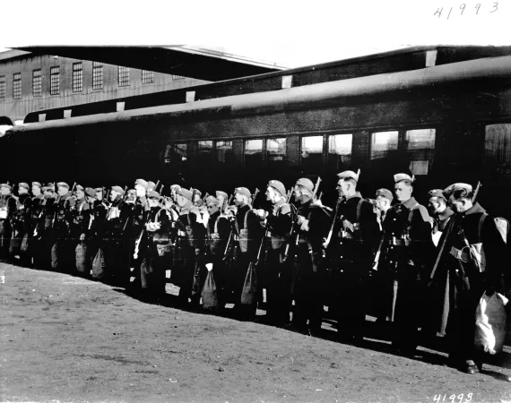 L’image est une photo noir et blanc montrant le premier contingent de soldats canadiens au garde-à-vous devant une voiture de train. 